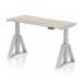 This picture features a sleek and modern dynamic sit stand and height adjustable desk. The desk measures 1400 x 600mm and has a grey oak top with cable ports and silver legs. The desk also includes a piste foot for added stability.