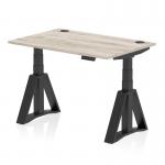 The photograph shows a sleek and modern office desk with a Grey Oak top and Black leg, equipped with cable ports for easy organization. The desk also features a Piste Foot for added stability and is height adjustable, allowing for both sitting and standing positions.