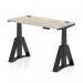 The photograph depicts a modern office desk with a grey oak top, black adjustable legs, and piste foot. Cable ports are visible for easy organization. The desk can transition between sitting and standing positions, providing a dynamic workspace.