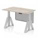 This dynamic sit stand and height adjustable desk features a modern grey oak top with silver legs and a silver steel modesty panel. The piste foot adds a sleek touch while providing stability. Perfect for any office setting.