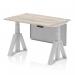 This photograph features a dynamic sit stand and height adjustable desk with a grey oak top, silver leg, and steel modesty panel. The desk also includes cable ports and a piste foot for added functionality.