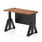 The photo shows a dynamic sit stand and height adjustable desk with a 1200 x 600mm walnut top, cable ports, and a black leg. The desk also features a black steel modesty panel and a piste foot for added stability.