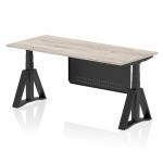 The photo shows a dynamic sit stand and height adjustable desk in an office setting. The desk has a grey oak top and black leg, with a black steel modesty panel and piste foot. The size of the desk is 1800 x 800mm. This versatile desk allows you to easily switch between sitting and standing positions while working.
