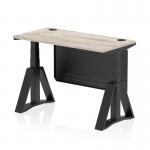 This photograph captures a sleek and modern office desk with a grey oak top and black steel legs. The desk is height adjustable and features cable ports, a modesty panel, and a piste foot for added stability. Its compact 1200 x 600mm size makes it perfect for any dynamic sit-stand work environment.
