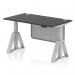 The photograph shows a sleek and modern office desk with a black top and silver legs. The desk has a dynamic sit stand feature, making it adjustable in height. It also has cable ports and a modesty panel with a silver steel finish. The piste foot adds stability to the overall design.