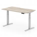 The image shows a stylish and modern grey oak top office desk with a scalloped edge, sitting on top of a sleek silver leg. The desk is equipped with an adjustable mechanism, allowing the user to switch between sitting and standing positions for a more dynamic work experience. Measuring 1400 x 800mm, this desk is perfect for any office space.