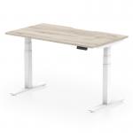 The photo shows a modern and sleek office desk with a grey oak top and white legs. The unique scalloped edge adds a touch of style. It is designed to be adjustable in both height and sitting positions, making it a functional choice for any workspace.