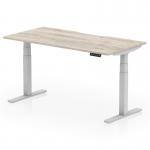 The photo shows a sleek, modern office desk with a scalloped edge grey oak top and silver legs. The desks height is adjustable, and it can be used for both sitting and standing positions. Its dimensions are 1600 x 800 mm, making it a versatile and functional addition to any workspace.