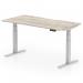 The photo shows a sleek, modern office desk with a scalloped edge grey oak top and silver legs. The desks height is adjustable, and it can be used for both sitting and standing positions. Its dimensions are 1600 x 800 mm, making it a versatile and functional addition to any workspace.