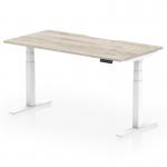 The picture shows a sleek and modern office desk with a grey oak top and white legs. The desk has a scalloped edge design and can be adjusted to different heights, making it suitable for both sitting and standing. It measures 1600 x 800mm and is perfect for a dynamic and adaptable workspace.