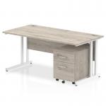 The photo shows a sleek and modern office desk with a grey oak top and white cantilever legs. The desk measures 1600 x 800mm and includes a 2-drawer mobile pedestal for storage. This dynamic furniture package is perfect for any professional workspace.