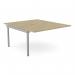 The photograph shows a sleek and modern office bench desk with add-on units. The desk features a scallop top and is 1400 x 800 in size, made with a stylish urban oak finish. It is designed for 2 people and includes a silver SLV panel for added functionality.