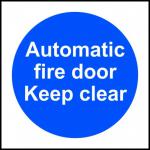 This photograph features a brightly colored industrial fire safety sign with the words Automatic Fire Door Keep Clear in bold letters. The sign is made of self-adhesive vinyl and measures 100mm x 100mm. It is designed to alert individuals to keep the area near the fire door clear at all times.