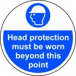 A photograph of this floor graphic shows a circular, anti-slip sign with the message Head Protection Must Be Worn Beyond This Point. The industrial design features bold white lettering on a black background and is laminated for durability.