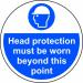 A photograph of this floor graphic shows a circular, anti-slip sign with the message Head Protection Must Be Worn Beyond This Point. The industrial design features bold white lettering on a black background and is laminated for durability.