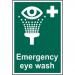 The picture shows a vibrant, eye-catching first aid sign featuring the words Emergency Eye Wash in bold lettering and a white symbol of an eye with water droplets. The sign is made of self-adhesive vinyl and measures 200mm x 300mm. Its striking colors and clear design make it easily visible and recognizable for quick access during emergencies.