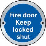 The picture shows a round satin anodised aluminium door disc with the words Fire Door Keep Locked Shut engraved on it. It is a Spectrum Industrial room sign used to indicate that the fire door should be kept locked at all times for safety.