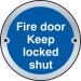 The picture shows a round satin anodised aluminium door disc with the words Fire Door Keep Locked Shut engraved on it. It is a Spectrum Industrial room sign used to indicate that the fire door should be kept locked at all times for safety.