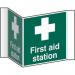 The photograph shows a vibrant Spectrum Industrial First Aid Signs sign, specifically a projection sign for a first aid station. It is made of 1.2mm recyclable PP and has a 200mm face. The bright and bold sign is designed to catch attention and clearly indicate the location of a first aid station.