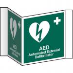This photo features a bright yellow Spectrum Industrial First Aid sign, with the words AED and First Aid in bold black letters. The sign is made of 1.2mm recyclable PP and has a 200mm face, making it easily visible. The image also shows that the sign is projected outward, making it easy to see from a distance.