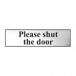 The photo shows a sleek, chrome-effect Spectrum Industrial Room Sign with the words Please Shut The Door in bold letters. The self-adhesive PVC material is 200mm x 50mm in size.