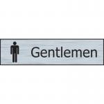 This photograph shows a Spectrum Industrial washroom sign labeled Gentlemen in a sleek stainless steel effect. The sign is made of self-adhesive PVC and measures 200mm x 50mm.