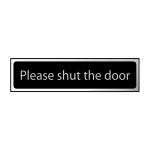 This photograph features a rectangular industrial room sign from Spectrum with the message Please shut the door printed in bold letters. The sign measures 200 x 50mm and has a blue background with white text. The CHR design adds a modern touch to the sign, making it suitable for various industrial settings.