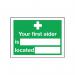 This photo features a rectangular First Aid sign with white text on a red background. The sign is self-adhesive and measures 150x200mm. The message reads Your First Aider Is with a blank space for customization. The sign includes the safety symbol E42AS and product code SR71214.
