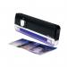 The photo shows a Safescan Hand Held UV Lamp 40H 130-0444 SSC33341, used for detecting counterfeit money. The compact device has a sleek black body and emits a strong purple light. It rests on top of a bundle of paper bills and appears to be in use, with someone holding the lamp over the money.